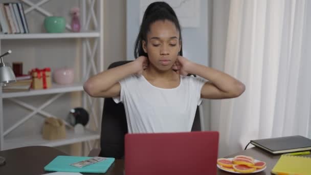 Retrato de plano medio de mujer afroamericana cansada y sobrecargada de trabajo que estira el espasmo del cuello sentada en la computadora portátil en la oficina en casa. Triste joven hermosa freelancer en el interior. Estilo de vida sedentario y atención médica. — Vídeos de Stock