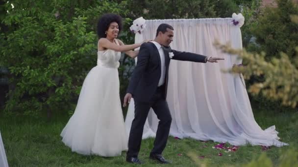 Divertido baile de alegres recién casados afroamericanos en el jardín de verano en el altar de la boda. Amplio plano de novia alegre en vestido y novio en traje bailando divirtiéndose al aire libre en ceremonia de matrimonio. — Vídeos de Stock