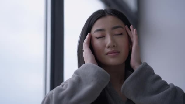 Portrait of exhausted tired Asian young woman rubbing temples in slow motion. Sad overworked overburdened beautiful slim millennial in bathrobe in home office at window indoors. Migraine and headache. — Stock Video