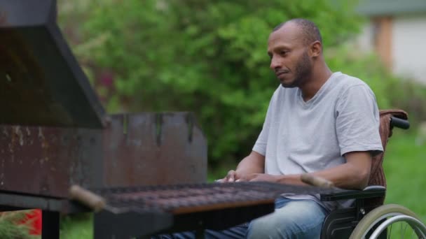 Medelskott porträtt av afroamerikanska handikappade man i rullstol sätta ved i grill i slow motion. Koncentrerad handikappad kille förbereder picknick utomhus på bakgården. — Stockvideo