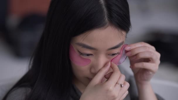 Close-up face of young slim Asian woman applying eye patches in slow motion. Headshot of beautiful charming gorgeous lady taking care of skin in bathroom indoors. Beauty and cosmetics concept. — Stock Video