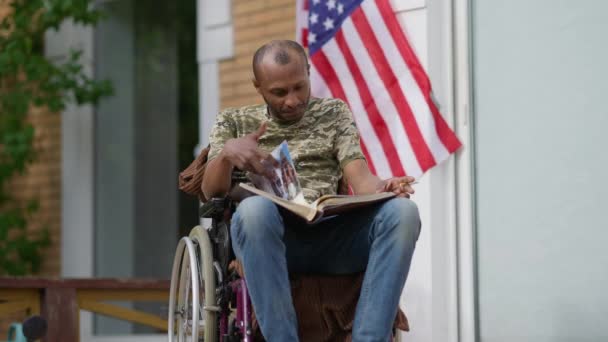Porträtt av handikappade afroamerikanska veteran minns minnen som tittar igenom fotoalbum sitter i rullstol på verandan med cigarett. Sorglig handikappad man på bakgården utomhus. — Stockvideo