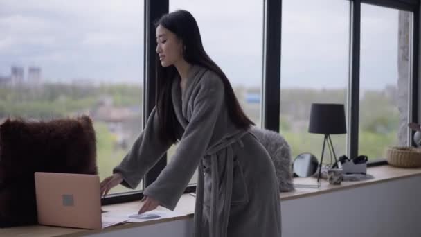 Portré vékony, magabiztos, gyönyörű ázsiai nőről, aki laptopot visz az ablakpárkányra, és reggel e-maileket néz. Bájos hölgy fürdőköpenyben dolgozik online otthoni irodában. — Stock videók