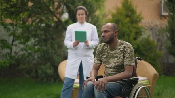 Грустный отчаявшийся инвалид в инвалидной коляске, смотрящий в сторону, как медсестра, стоящая на заднем плане. Портрет безнадежного афроамериканского военного пациента и кавказского сиделки в доме престарелых. — стоковое видео