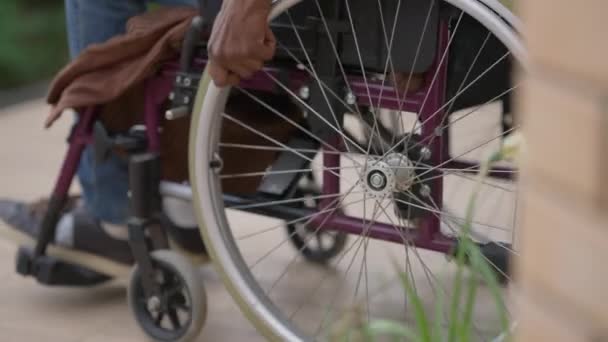 La cámara se mueve desde las ruedas en silla de ruedas hasta el hombre afroamericano discapacitado rodando al aire libre en el porche. Triste hombre adulto parapléjico a caballo dispositivo operado manualmente en el día de primavera de verano. — Vídeos de Stock