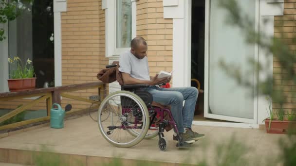Απορροφημένος ανάπηρος Αφροαμερικανός διαβάζει βιβλίο καθισμένος σε αναπηρική καρέκλα στη βεράντα. Ευρεία βολή των ατόμων με ειδικές ανάγκες έξυπνο ανάπηρος απολαμβάνοντας χόμπι σε εξωτερικούς χώρους για ηλιόλουστη μέρα του καλοκαιριού στην πίσω αυλή. — Αρχείο Βίντεο