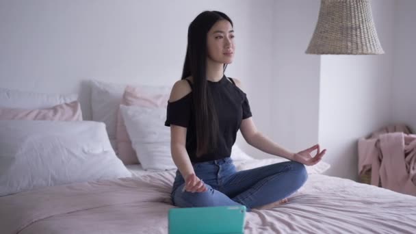 Brett skott av lugn säker asiatisk kvinna sitter i lotus poserar på sängen leende med tablett liggande åt sidan. Porträtt av smal vacker frilansare som gör avslappningsövning på hemmakontoret. Yoga och affärer. — Stockvideo