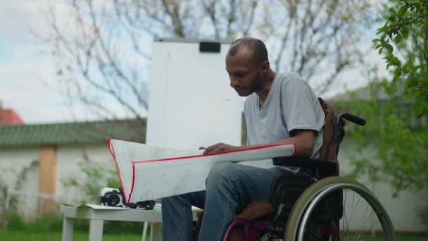Зосереджений на талановитому ландшафті, він аналізує креслення на відкритому повітрі. Портрет серйозного інтелігентного афроамериканського чоловіка в кріслі на колесах, який переглядає папери на відкритому повітрі. Розум і інвалідність. — стокове відео