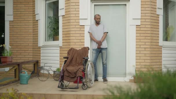 Ευρύ πορτραίτο ενός όμορφου Αφροαμερικανού που στέκεται στη βεράντα με αναπηρική καρέκλα και περιμένει. Ενήλικες νοσοκόμοι ή συγγενείς που φροντίζουν άτομα με ειδικές ανάγκες στο σπίτι ή στο γηροκομείο. — Αρχείο Βίντεο