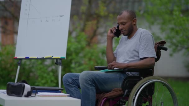Side view of confident disabled expert in wheelchair discussing business idea on the phone sitting in summer garden outdoors. Serious professional African American handicapped man talking. — Stock Video