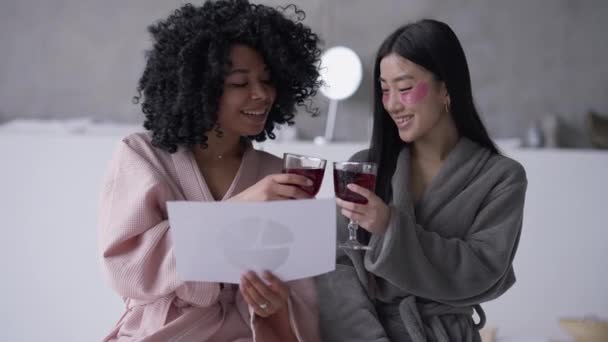 Lachende succesvolle jonge vrouwelijke startupers klinkende sap glazen drinken gezonde drank analyseren van de winstgevendheid grafiek. Portret van positieve Afro-Amerikaanse en Aziatische vrouwen binnen. — Stockvideo