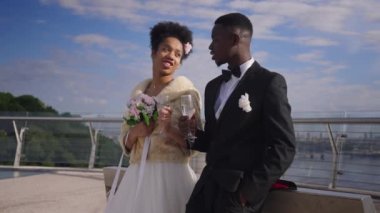 Mutlu gelin ve damat kadehlerini tokuşturarak dışarıda şampanya içiyorlar. Gülümseyen Afro-Amerikalı kadın ve erkek, yaz günü köprüde evliliği kutluyorlar. Neşe ve evlilik.