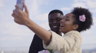 Neşeli, güler yüzlü bir çift yaz günü açık havada düğün töreninde selfie çekiyor. Mutlu genç Afro-Amerikan güzel gelin ve yakışıklı damat evleniyor akıllı telefondan fotoğraf çekiliyor..