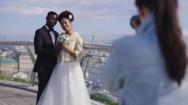Mutlu Afrikalı Amerikalı yeni evliler, güneşli yaz gününde briçte fotoğrafçıya poz veriyor. Gülümseyen zarif gelin ve damat düğün günü dışarıda fotoğraf çekiyorlar. Yavaş çekim.