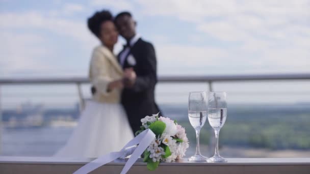 Copas de champán y ramo de novia tumbados en el banco con felices recién casados afroamericanos bailando en cámara lenta al fondo. Amar novia y novio disfrutando de la boda al aire libre en el puente. — Vídeos de Stock