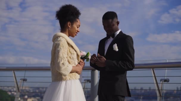 Vista lateral de tiro medio de novio confiado poniendo anillo de bodas en el dedo de la novia sonriente al aire libre. Feliz pareja afro-americana que se casa en bridge. Movimiento lento. — Vídeos de Stock