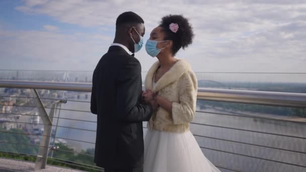 Pareja afro-americana cariñosa positiva en la cara de Covid-19 máscaras frotando narices cogidas de la mano de pie en el puente. Novia feliz y novio disfrutando de la boda al aire libre en la pandemia de coronavirus. — Vídeos de Stock