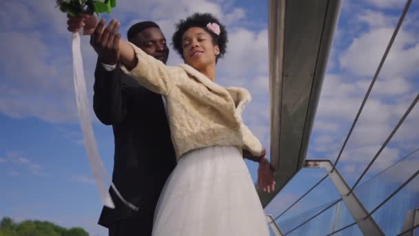 Botten vinkel syn på glad leende par nygifta höjer händerna imitera Titanic scen med sommar himmel i bakgrunden. Spännande tillgiven afroamerikansk brud och brudgum njuter av äktenskap. — Stockvideo
