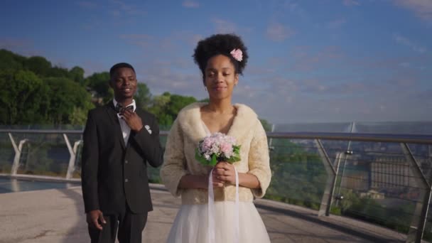 Porträtt av smal vacker afroamerikansk kvinna i brudklänning med brudbukett tittar på kameran ler som man i elegant kostym stående bakgrund. Glad brud poserar med brudgum utomhus. — Stockvideo