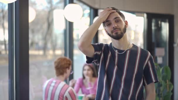 Zelfverzekerde jonge getatoeëerde man die in slow motion haar aanraakt en glimlachend naar de camera kijkt. Portret van een blanke brunette met bruine ogen die binnen poseert in een retro café. Leefstijl en vertrouwen in de jaren tachtig. — Stockvideo