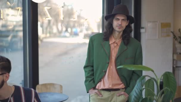 Confiado dandy de 1980 caminando en cámara lenta mirando a la gente sentada en la cafetería vintage. Retrato de hombre masculino caucásico con sombrero y chaqueta de terciopelo paseando en el interior. Concepto de estilo 1990. — Vídeo de stock