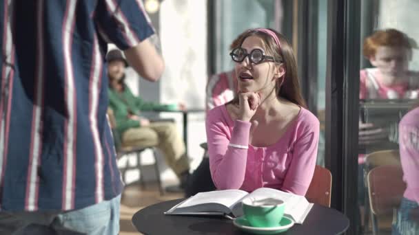 Porträtt av retro nörd kvinna sitter vid bordet i vintage café med bok tittar på mannen passerar. Pinsamt ledsen vit dam som väntar på blindträff inomhus på 1980-talet. Livsstilskoncept. — Stockvideo