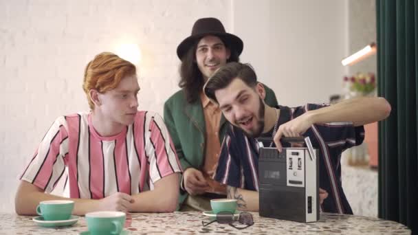 Smiling man winking turning on retro tape recorder and group of cheerful male friends dancing sitting at table in vintage coffee bar. Handsome young Caucasian men having fun in 1980s indoors. — Stock Video