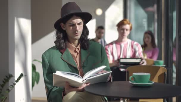 Joven hombre retro centrado en el libro de lectura sombrero sentado en la mesa en la cafetería vintage. Retrato de inteligente inteligente guapo confiado Caucásico chico disfrutando de hobby en 1980 en interiores. Estilo de vida. — Vídeo de stock