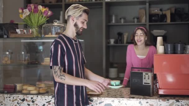 Getatoeëerde bebaarde mannelijke klant die koffiebeker neemt van vrouwelijke barista in vintage koffieschop, kijkend naar camera glimlachend ruikend en smakelijk drankje drinkend. 1980 vrijetijdsbesteding en levensstijl. — Stockvideo