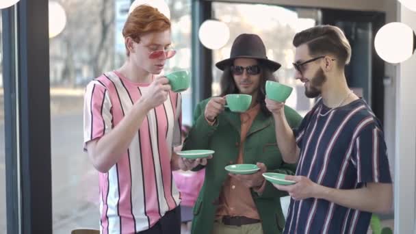 Drie positieve jonge Kaukasische retro mannen in een zonnebril die koffie drinken en glimlachen terwijl ze naar de camera kijken. Portret van gelukkige ontspannen vrienden rusten in koffiebar in de jaren 1980 1990 poseren. — Stockvideo