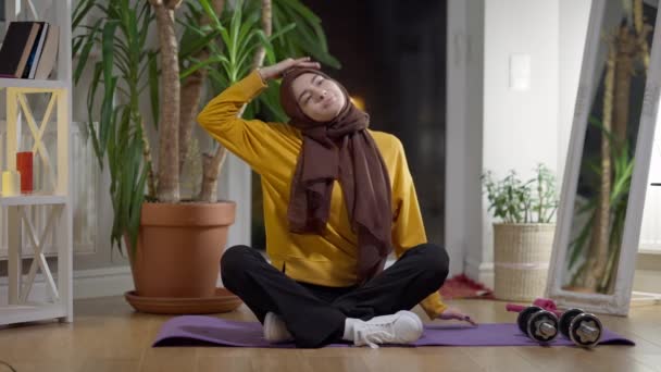 Amplia vista frontal de la mujer en forma en el hijab calentando los músculos sentados en el entrenamiento de la esterilla de ejercicio en casa por la noche. Retrato de una deportista delgada y hermosa ejercitándose en interiores. — Vídeos de Stock
