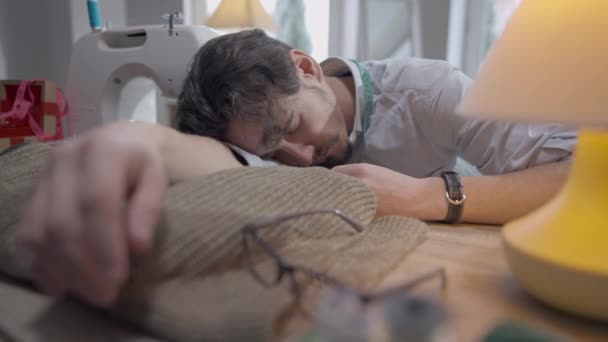 Joven sastre agotado durmiendo en la mesa en el estudio en el interior. Retrato del hombre caucásico guapo y cansado quemado con los ojos cerrados durmiendo en el taller sobre la ropa. Cansancio y creatividad. — Vídeos de Stock