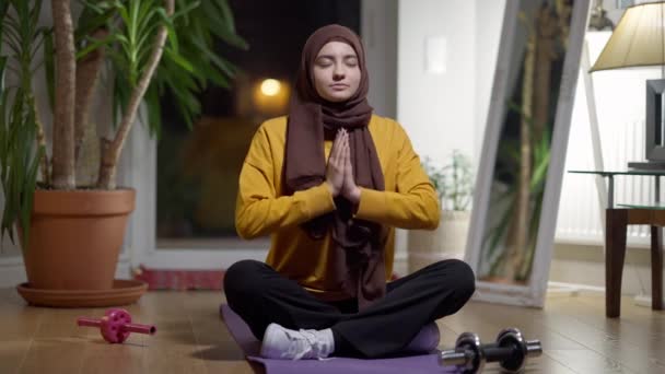 Amplia toma de confiada mujer de Oriente Medio meditando en pose de loto con los ojos cerrados. Retrato de una hermosa deportista delgada en hijab en casa por la noche. Concepto de deporte y tranquilidad. — Vídeos de Stock