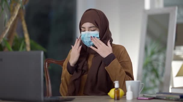 ヒジャーブで若いイスラム教徒の女性の中出しと鼻スプレーを示すホームオフィスのくしゃみでコヴィド顔マスク会議.隔離中のコロナウイルス病の症状を持つ中東のフリーランサー — ストック動画