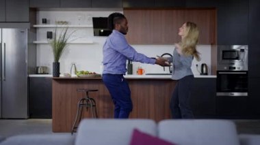 Arka planda mutfak ve oturma odasında yavaş çekimde dans eden neşeli çiftin geniş bir görüntüsü. Afro-Amerikan erkeği ve beyaz kadını mutlu bir şekilde sevin. Hafta sonunun tadını çıkarın..