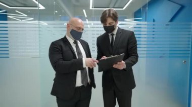 Orta yaşlı iş ortağıyla ofis koridorunda duran beyaz, zarif genç bir adamın portresi. Covid maskeli başarılı iş adamları koridorda anlaşma yapıyor..