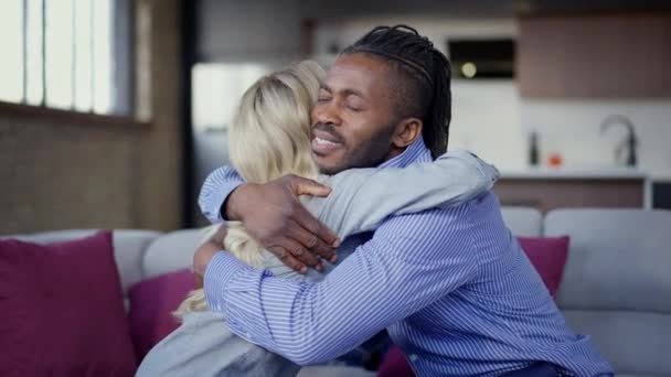 Vista lateral de amante interracial feliz pareja abrazándose en cámara lenta sentado en el sofá en la sala de estar. Sonriente esposa caucásica y esposo afroamericano se reúnen en casa por la noche. — Vídeos de Stock