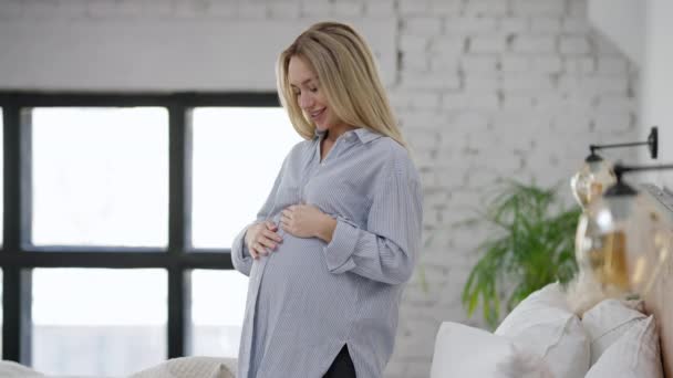 Sidovy av lycklig gravid kvinna pratar med ofödda fostret smekande mage. Glada leende vacker blond vit ung förväntansfull står i sovrum hemma. Graviditet och livsstil. — Stockvideo