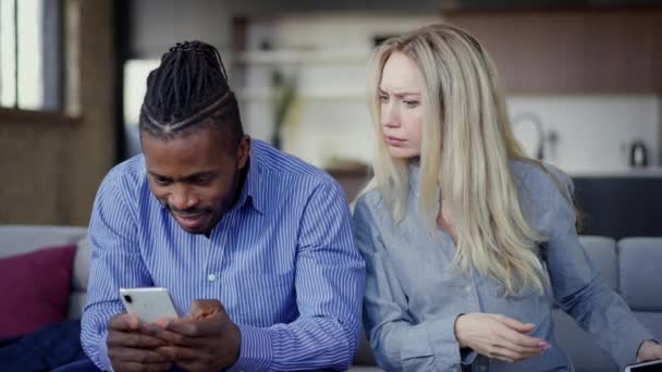Jealous Caucasian wife looking at smartphone of African American husband talking and gesturing. Envious young beautiful woman arguing with unfaithful handsome husband messaging online in social media. — Stock video