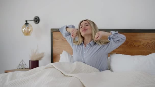 Happy Caucasian woman stretching in bed in the morning at home. Portrait of joyful smiling beautiful lady waking up in bedroom looking at camera. Leisure and beauty concept. — Stock video