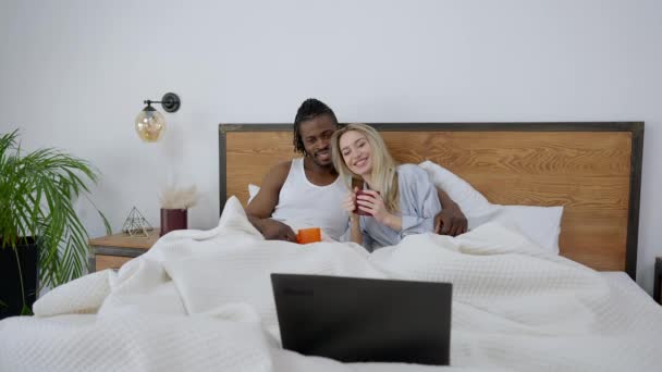 Weitwinkelporträt eines glücklich verliebten gemischtrassigen Paares, das abends im Bett liegt und auf dem Laptop Komödienfilme anschaut. Entspannt lachende Kaukasierin und Afroamerikaner genießen Film. — Stockvideo