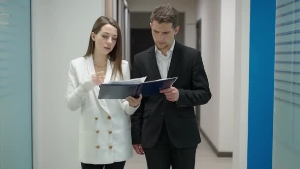 Busy young man and woman walking in office corridor discussing project analyzing documents as coworkers passing. Portrait of concentrated colleagues talking and strolling in hallway. Live camera. — Stock video