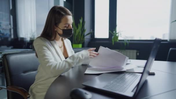 Coronavirus 'lu beyaz kadın portresi ofisteki dizüstü bilgisayarın klavyesinde yazan belgeleri analiz ederken maske takıyor. Kapalı alanda Covid-19 salgınında çalışan konsantre kadın çalışan.. — Stok video