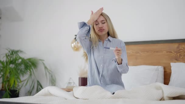 Een blanke vrouw met een positieve zwangerschapstest die een gebaar maakt. Portret van onwillige zwangere jonge mooie dame zitten op bed met versufte gezichtsuitdrukking. — Stockvideo