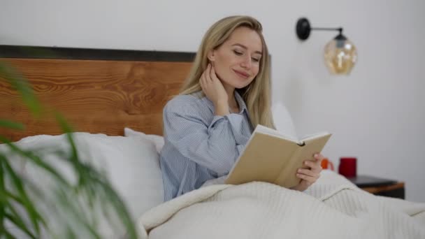 Ung charmig intelligent vit kvinna läser bok sitter i sängen leende. Porträtt av smart säker vacker tusenårig njuter hobby i sömnen i sovrummet. Livsstil och glädje. — Stockvideo