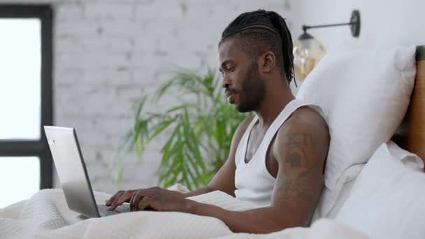 Vista lateral de concentrado bonito tatuado afro-americano homem digitando no teclado laptop em câmera lenta sentado na cama. Freelancer confiante focado navegando mensagens de Internet on-line pela manhã. — Vídeo de Stock