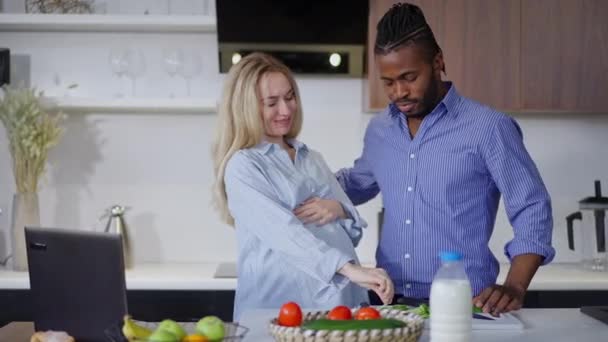 Femeie gravidă caucaziană care degustă ingrediente organice pentru salată sănătoasă ca bărbat afro-american îmbrățișând soțul vorbind în mișcare lentă. Portretul cuplului interrasial gătit micul dejun acasă . — Videoclip de stoc