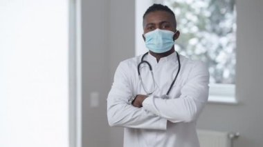 Medikal üniformalı kendinden emin Afro-Amerikan bir adamın orta boy portresi ve kamera karşısında el ele tutuşan koronavirüs yüz maskesi. Covid-19 'da hastanede poz veren profesyonel bir doktor..