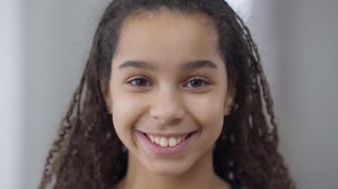 Afro-Amerikalı güzel, kahverengi gözlü ve gülümseyen kameraya gülümseyen dişlek bir genç kızın vesikalığı. Kapalı mekanda poz veren neşeli güzel gençlerin yakın çekimi. Neşe kavramı.