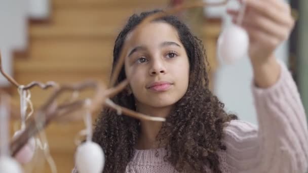 Портрет прекрасної афроамериканської дівчини-підлітка, яка прикрашає філіал іграшковими яйцями, що посміхаються, дивлячись на камеру. Радісний щасливий підліток, що подає себе вдома на Великдень. Святкова концепція. — стокове відео
